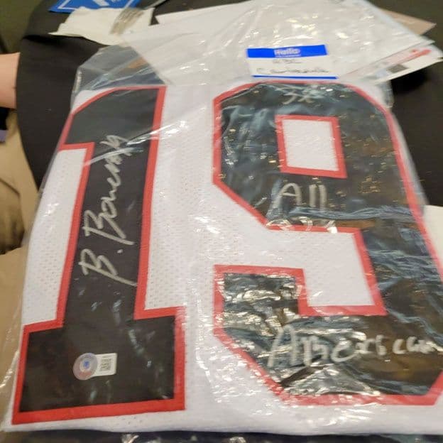 A sports jersey with the number 19, featuring multiple signatures in white, is displayed in protective plastic on a dark surface.