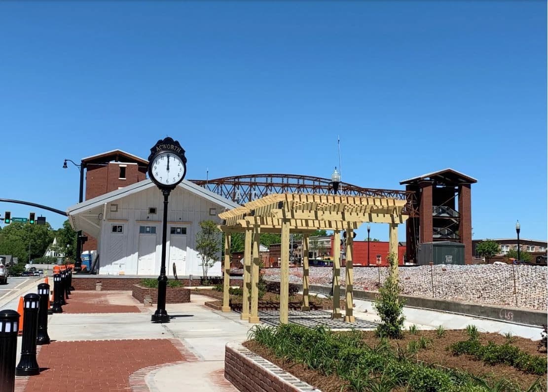 Historic town square under clear blue sky with a vintage clock, brick pathways, a wooden pergola, and surrounding greenery. Tranquil and inviting scene.