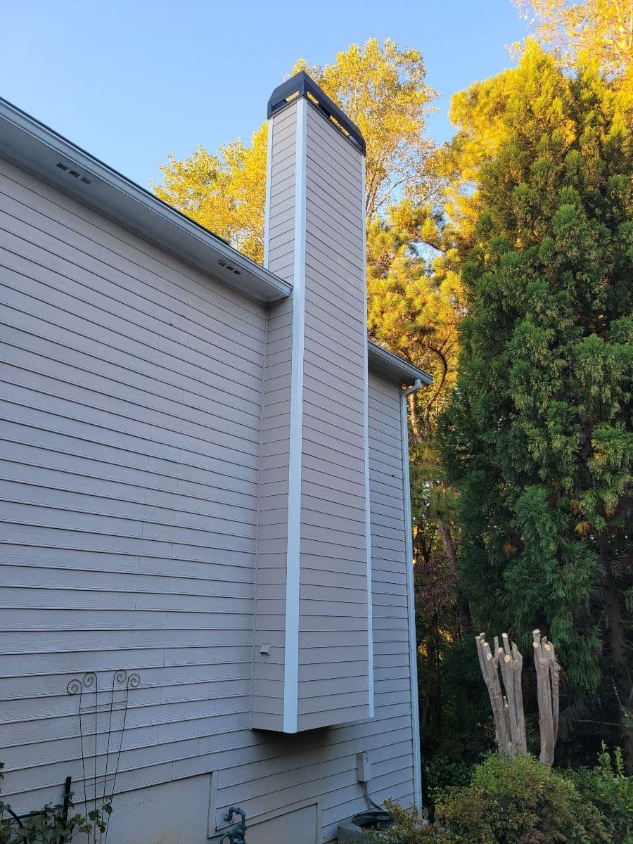 A tall, rectangular chimney stretches up the side of a beige house, surrounded by lush green trees under a clear blue sky at sunset.