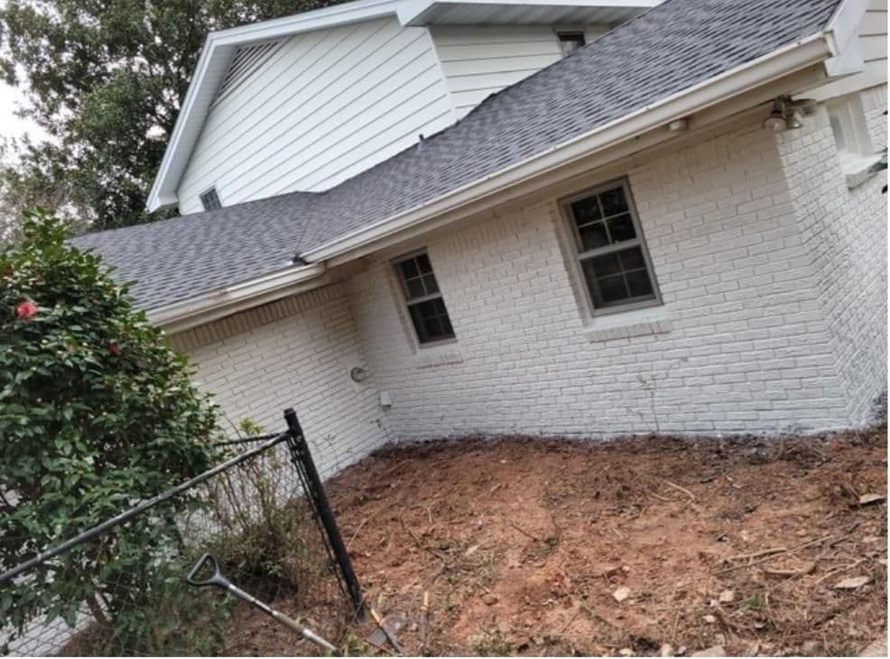 Side view of a white brick house with a sloped grey roof. The yard is bare soil, with a shovel leaning on a black fence. A bush is to the left. Calm atmosphere.