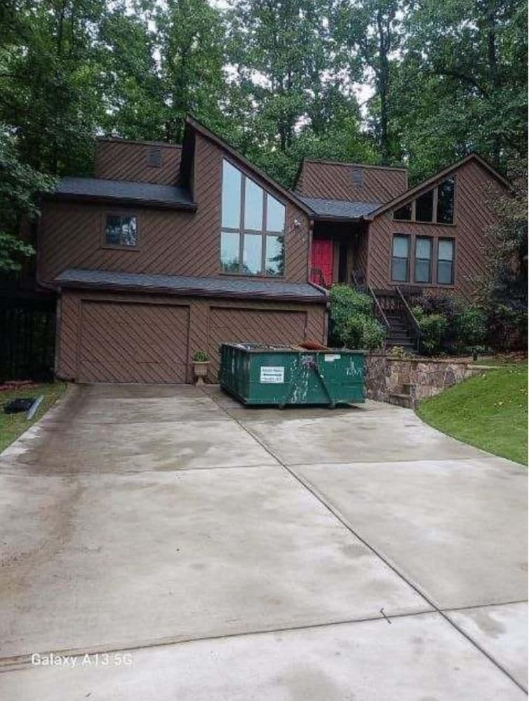 Modern brown house with large windows, surrounded by trees. A green dumpster is placed at the end of the smooth concrete driveway. Overcast sky.