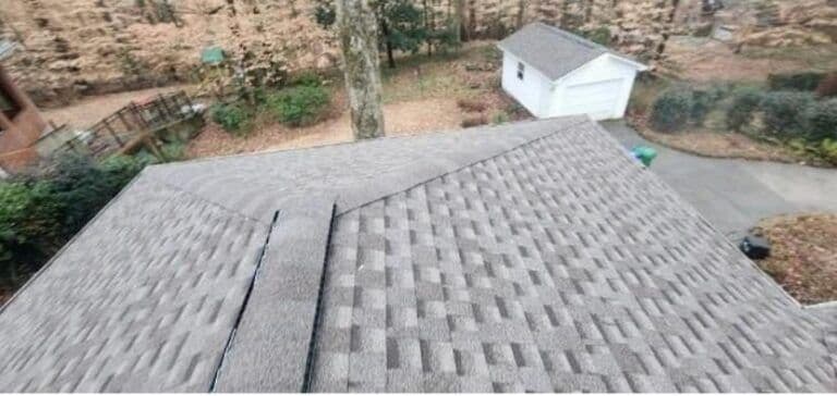 Aerial view of a gray shingled rooftop with geometric patterns. Background shows a detached white garage, driveway, and trees with autumn foliage. Calm suburban setting.