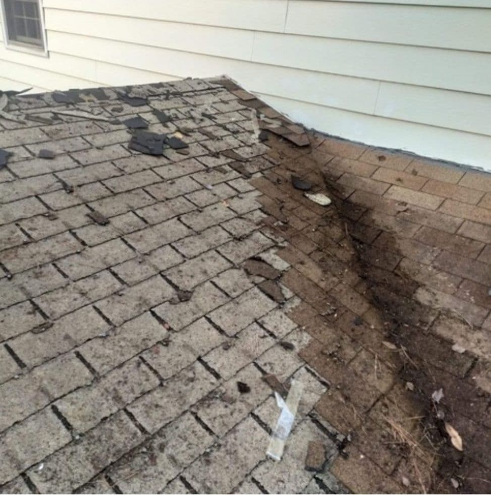 Damaged roof with missing and cracked shingles and exposed underlayer by a beige siding wall, suggesting deterioration and neglect.