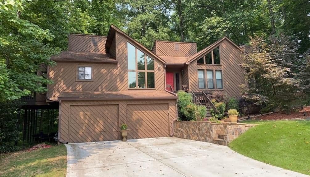 A modern brown house with large windows, a red front door, and a two-car garage. Surrounded by lush trees and a manicured lawn, it conveys tranquility.