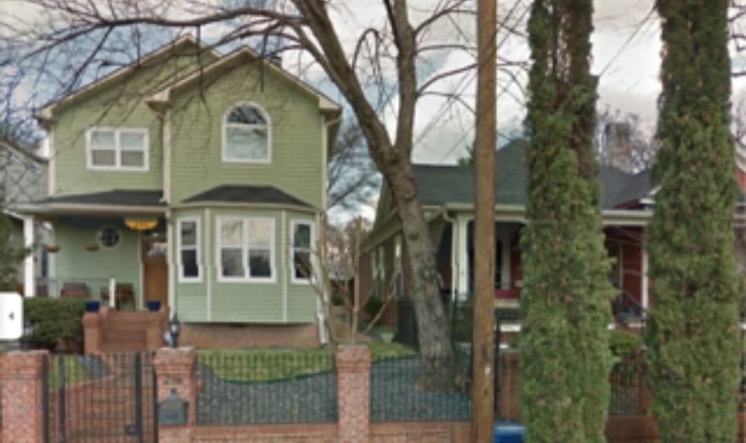 A light green two-story house with white trim sits behind a brick wall. Trees and a neighboring home are visible, conveying a suburban neighborhood scene.