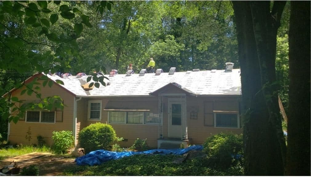 A house with roof work in progress, surrounded by lush trees. Several workers are on the roof with materials, conveying a sense of productivity and renovation.