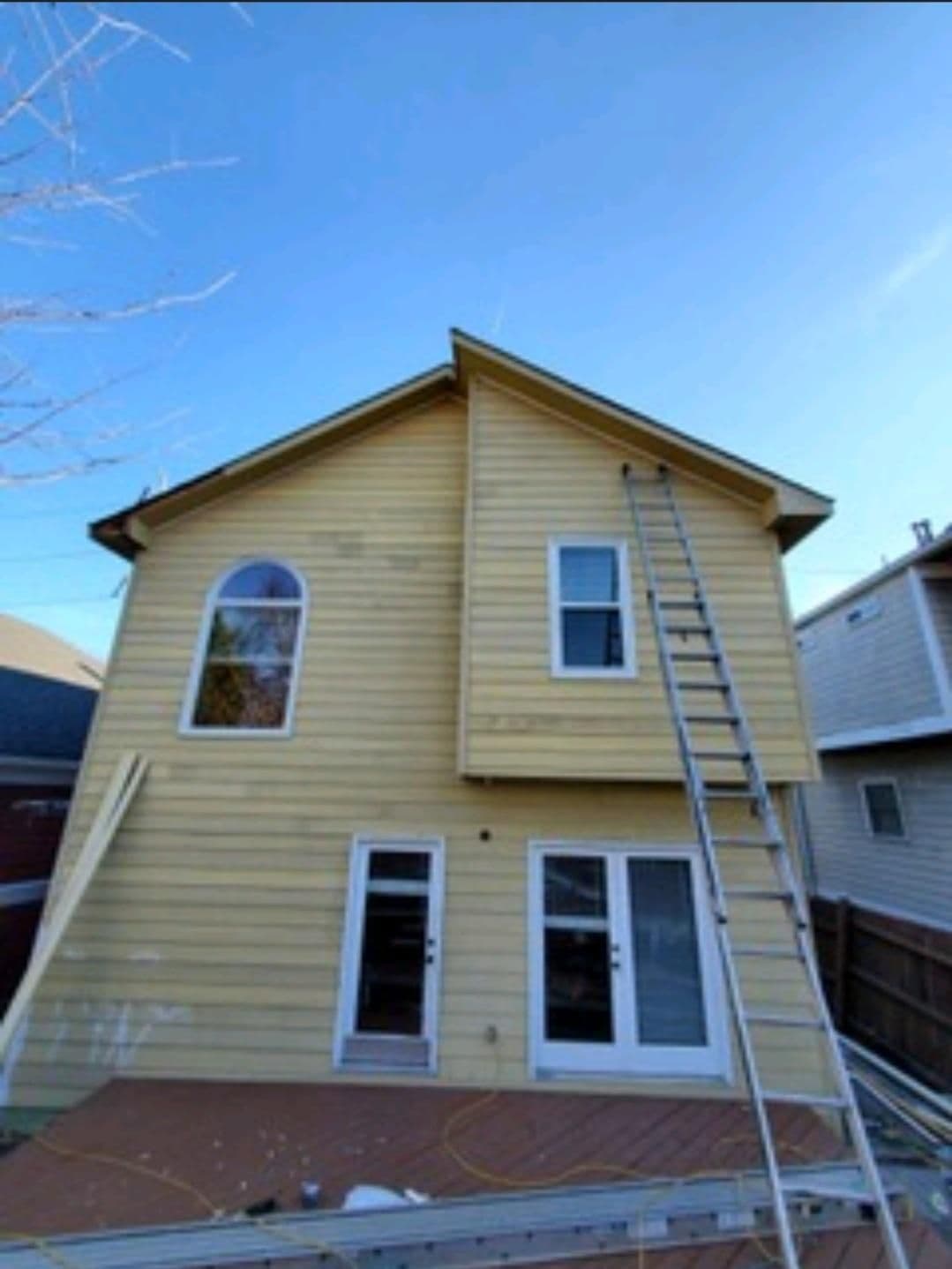 Two-story yellow house under clear blue sky; a tall ladder leans against it, suggesting ongoing work. Glass doors and arched window visible.