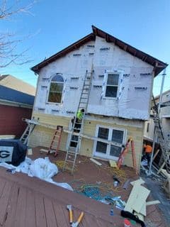 A house under renovation with ladders and construction materials. Workers apply Tyvek wrap on the upper walls, tools scattered on the wooden deck.