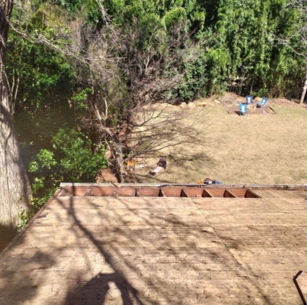 View from a roof showing a shadow of a person taking the photo. Below, a grassy backyard with trees and scattered blue buckets conveys a tranquil outdoor setting.