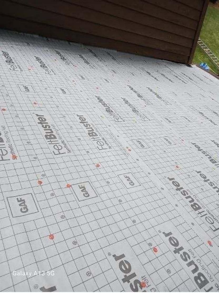 Close-up of a roof covered with GAF FeltBuster underlayment. The surface has a grid pattern with markings, attached with orange fasteners. Wooden siding visible.
