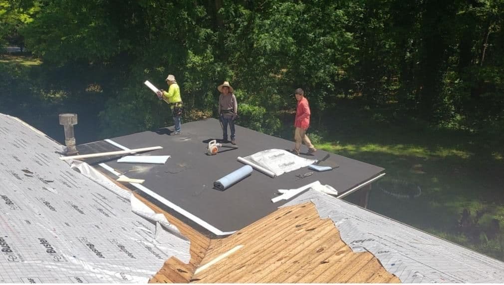 Three workers stand on a roof under construction, surrounded by various tools and materials. The background is lush with green trees, and the scene conveys teamwork and diligence.