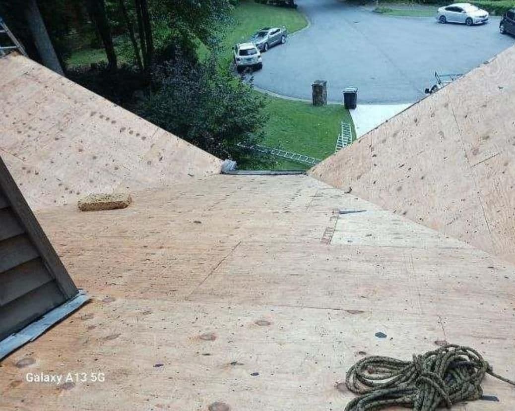 View from a steep plywood roof overlooking a suburban street. A rope and sponge lie on the roof. Cars and greenery are in the background.