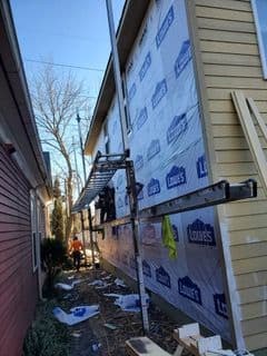 House under construction with worker on scaffolding. Exterior wall covered in Lowe's wrap and partial siding. Clear blue sky, sunny day.