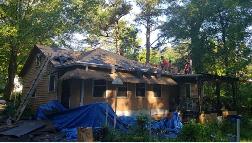 Two workers on a partially shingled roof repair a house surrounded by trees. A ladder leans against the house, and blue tarps cover the ground.