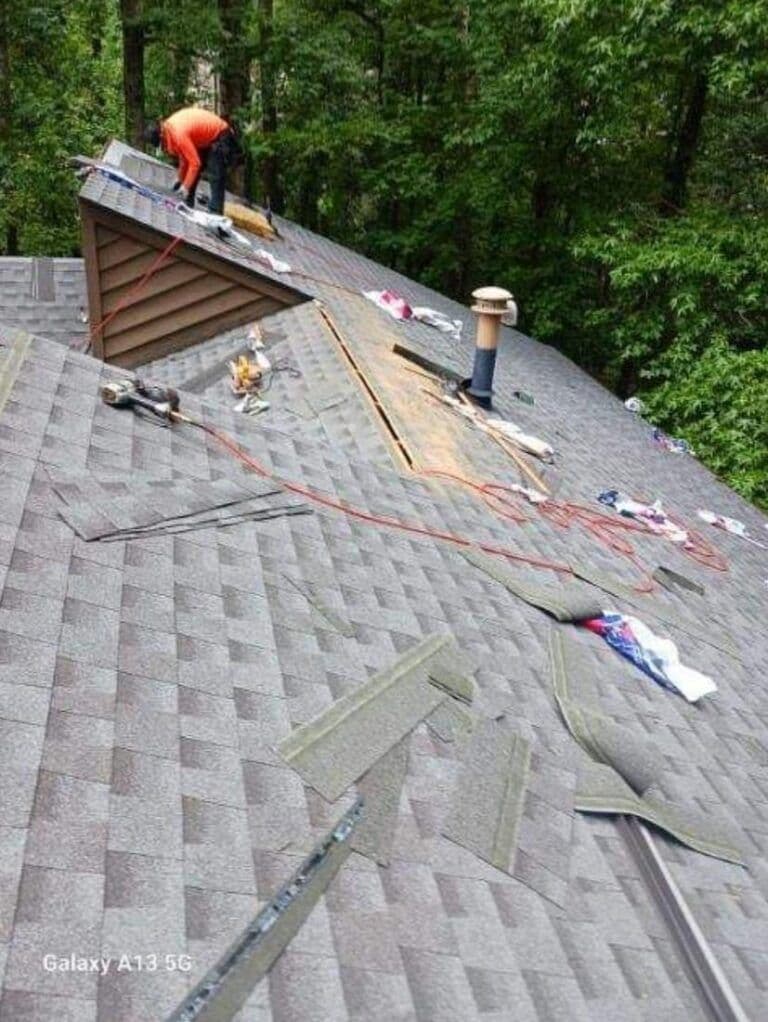 A worker in an orange shirt repairs a sloped roof with shingles. Tools and materials are scattered. Dense green trees surround the scene.