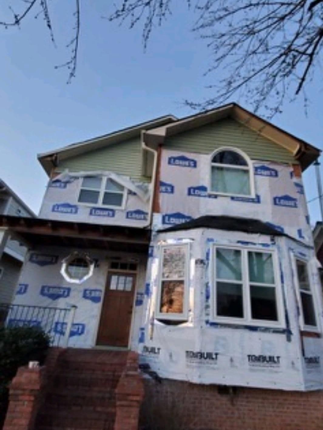 Two-story house under renovation, wrapped in Lowe's branded building paper. Exposed brick steps lead to a wooden door. Clear sky visible above.