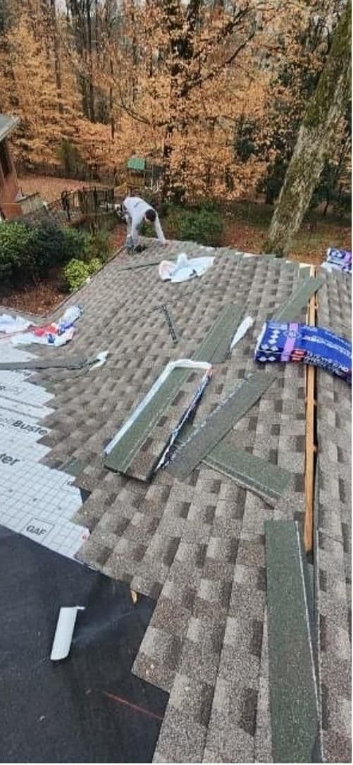 A worker is installing gray shingles on a house roof surrounded by autumn trees. Tools and roofing materials are scattered, creating a busy scene.