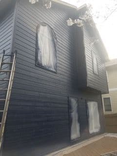 A two-story house is partially painted dark blue with windows covered for protection. A ladder leans against the wall, suggesting ongoing work.