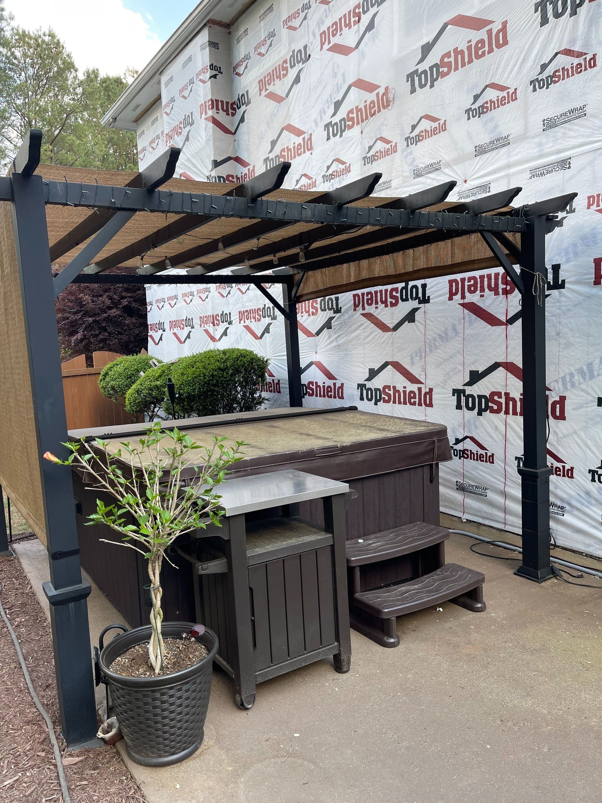A hot tub under a black pergola in a backyard. There's a small table and potted plant in front. The house is covered with TopShield wrap.