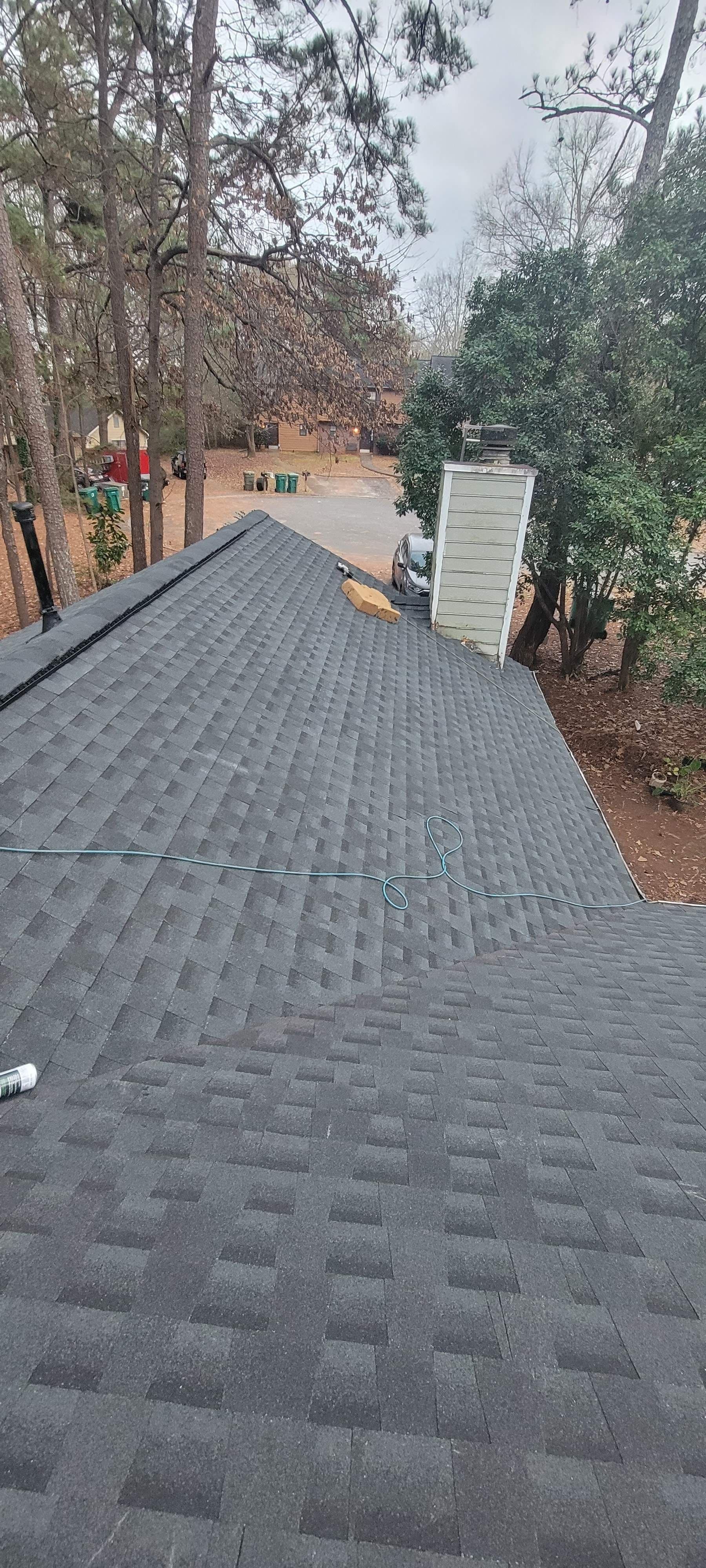 Gray shingled roof surrounded by trees and a cloudy sky. A ladder leans against the house, while a garden hose lies on the roof, suggesting maintenance work.
