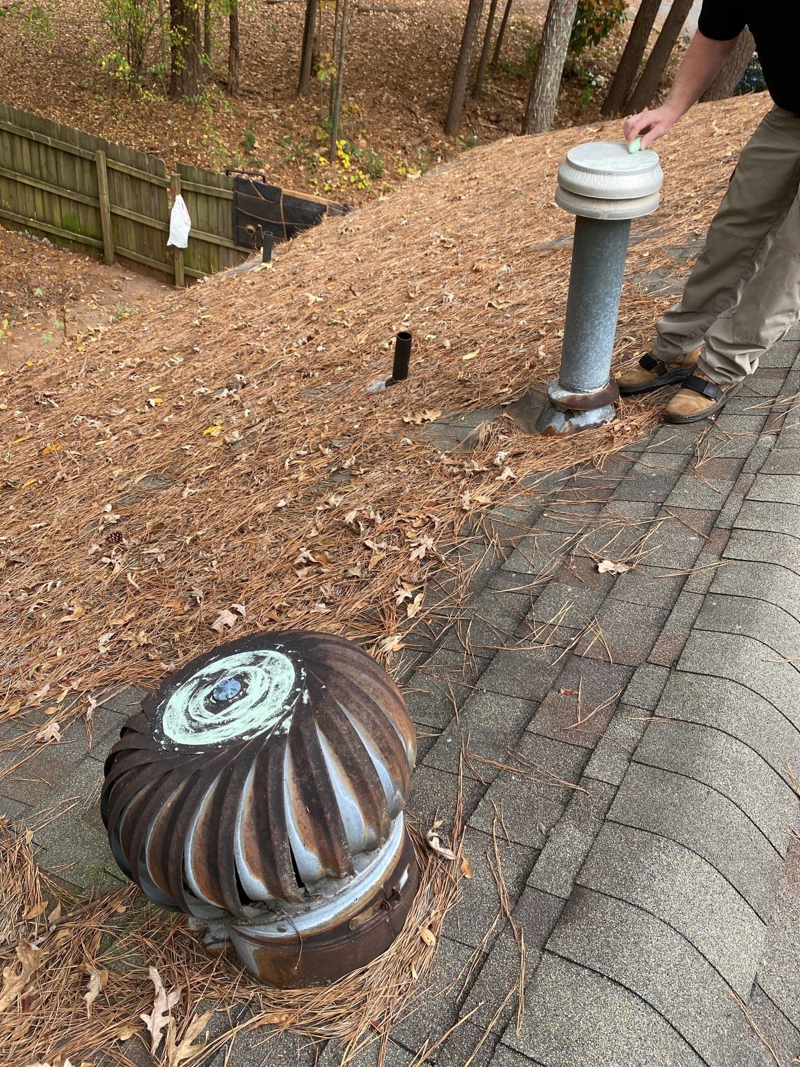 A roof with two metal turbine vents partially covered in pine needles. A person adjusts one vent, surrounded by trees and a wooden fence.