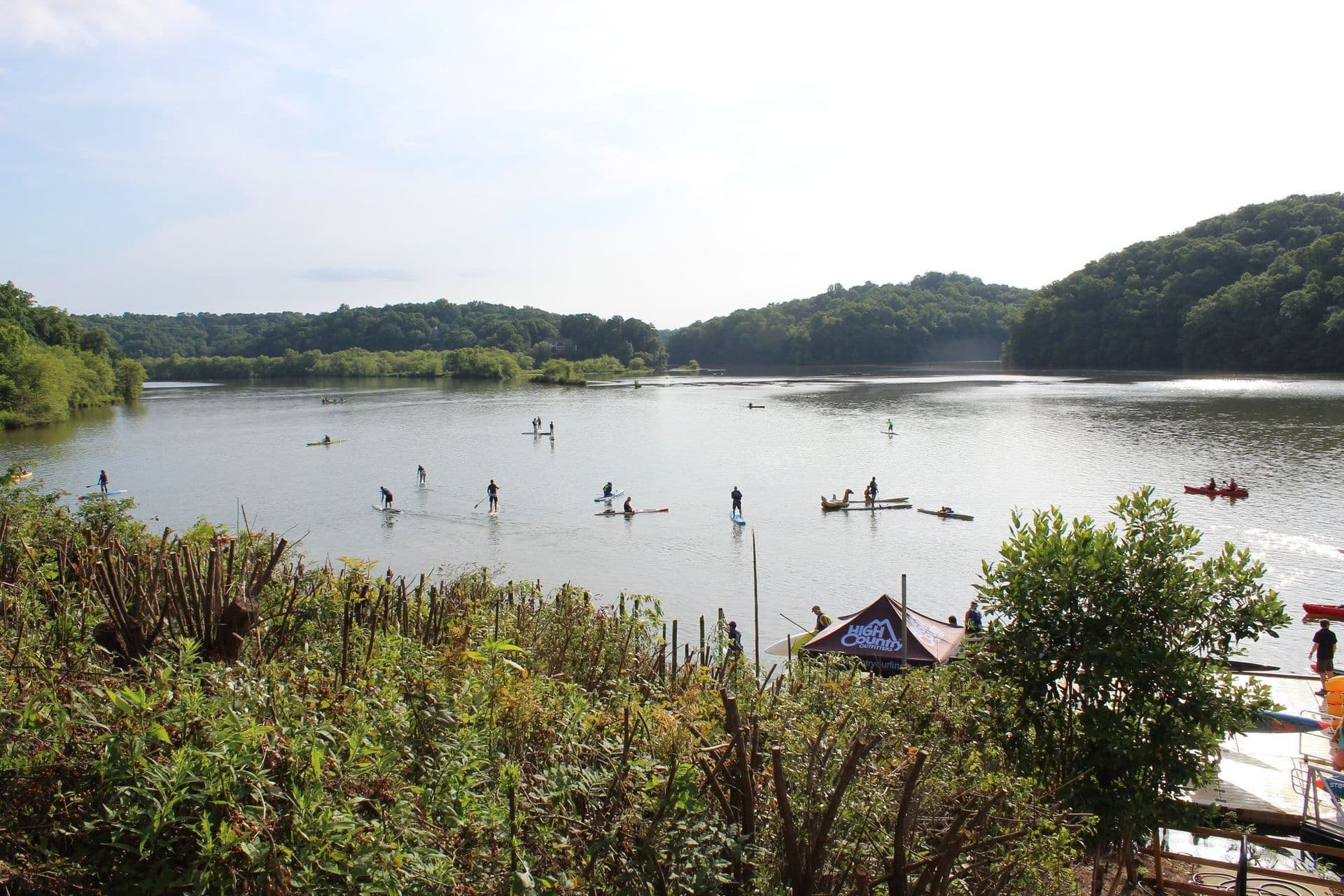 A serene lake scene with people paddleboarding and kayaking under a bright sky. Dense green foliage frames the scene, conveying a peaceful atmosphere.