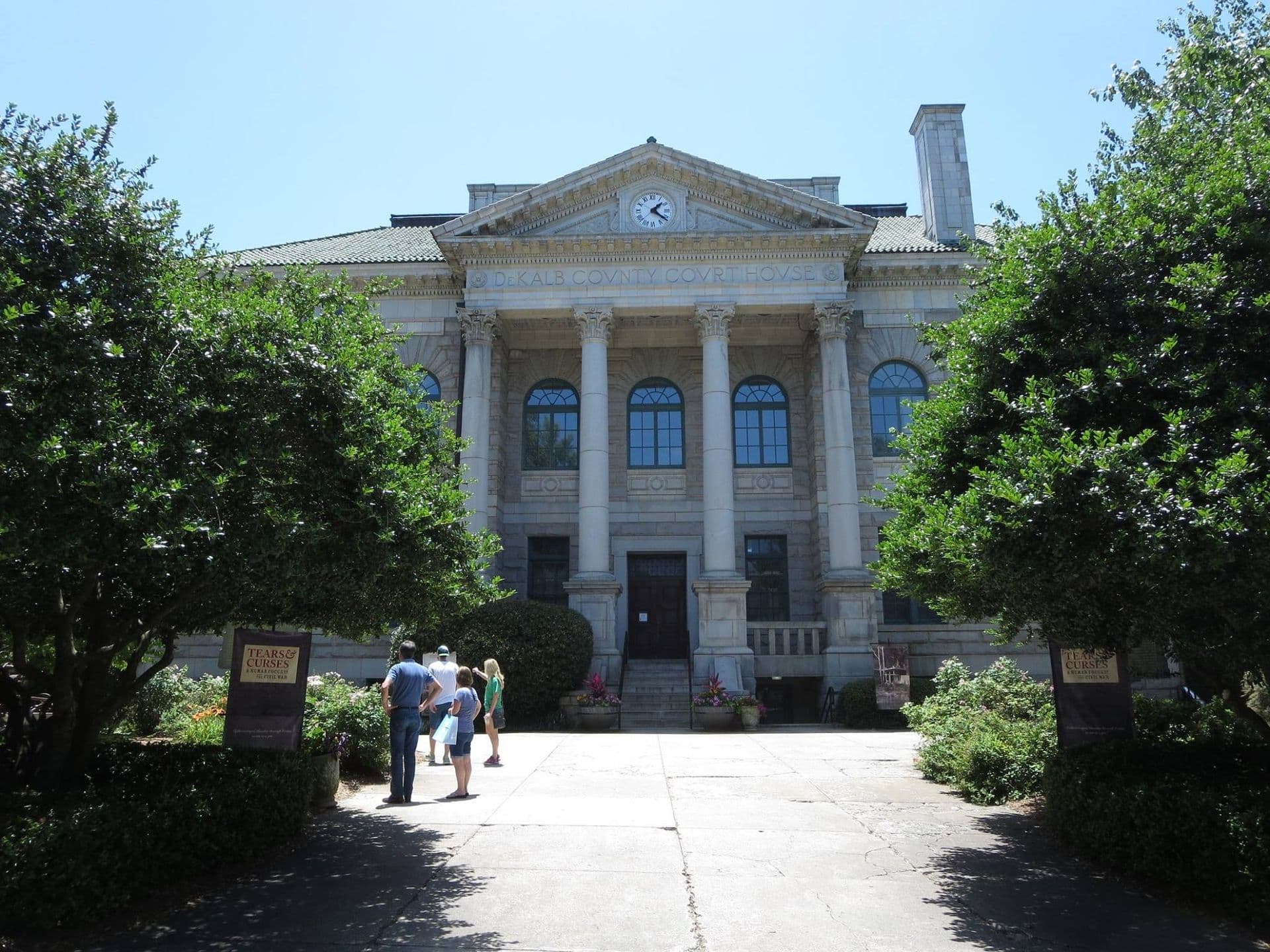 A historic stone building with four columns, arched windows, and a clock above the entrance. People stand outside, surrounded by green trees and clear blue skies.