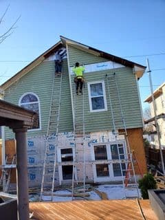 Two workers on ladders install siding on a house's upper level. The lower half is wrapped in protective material. Clear blue sky suggests sunny weather.