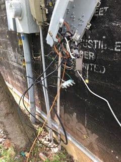 Messy cluster of exposed electrical wires and cables hanging from a damaged utility box on a brick wall, suggesting disarray and potential hazard.