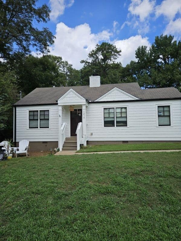 A small, white, single-story house with a gray roof sits surrounded by lush greenery and trees. The sky is blue with scattered clouds, conveying a peaceful suburban scene.