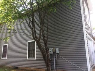 A gray house exterior with two windows, an electrical meter, and a tree in the foreground. Sunlight casts shadows, conveying a calm, suburban feel.