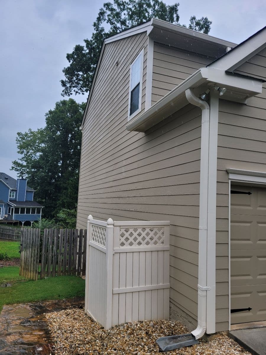 Side view of a beige house with rain, featuring a drainpipe and lattice-topped privacy fence. Wet pebble and stone path in the foreground; trees in the background.