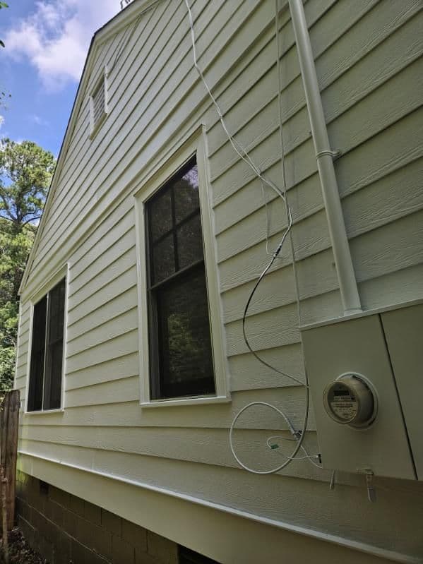 Side view of a house with light-colored siding. Two dark-framed windows and an electric meter are visible. Overhead wires connect to the meter.