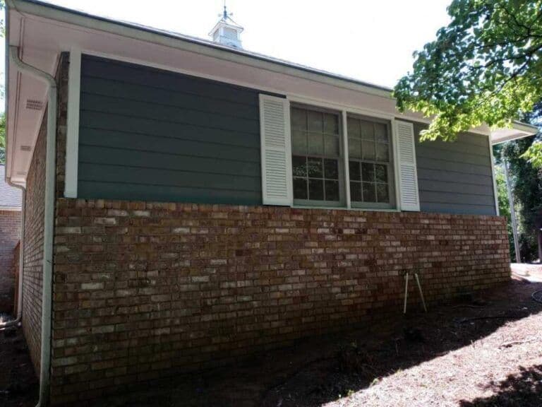 A house exterior with a mix of brick and blue siding. Large windows with white shutters, partly shaded by a nearby tree, creating a calm suburban feel.