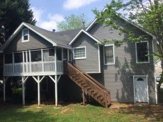 Elevated gray house with a screened porch and wooden staircase. Surrounded by greenery, the scene is peaceful and sunny, evoking a cozy suburban feel.