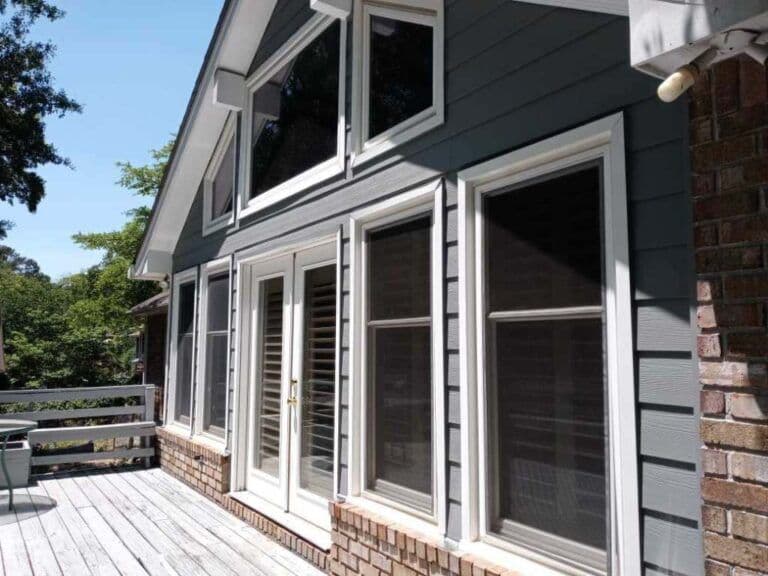 A sunlit gray house exterior features large windows and French doors on a porch. The bright, inviting scene includes leafy trees in the background.