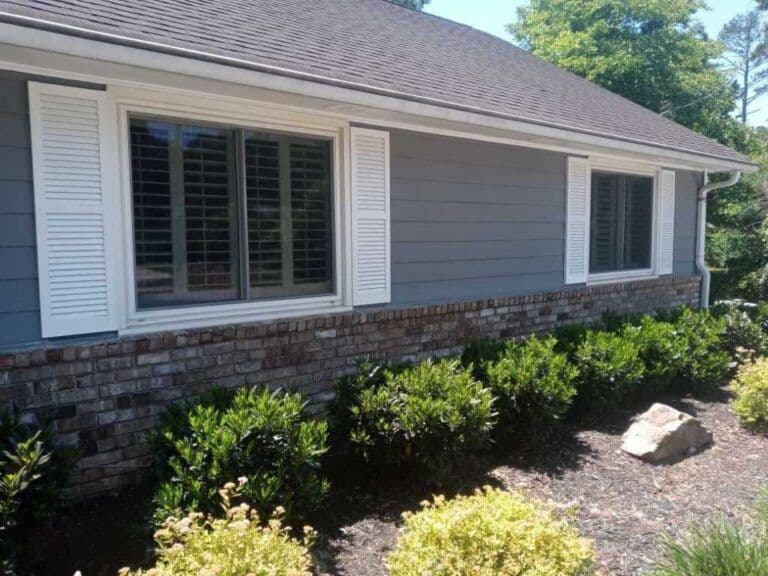 Side view of a house with blue-gray siding and brick foundation, featuring two windows with white shutters. Bushes and a rock in the landscaped garden. Bright, sunny day.