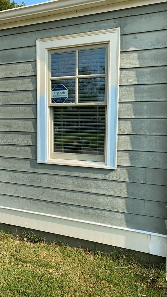 A light blue-gray wooden wall with a white-trimmed window displaying a "SimpliSafe" security sign. Sunlit grass at the bottom conveys a calm, secure setting.