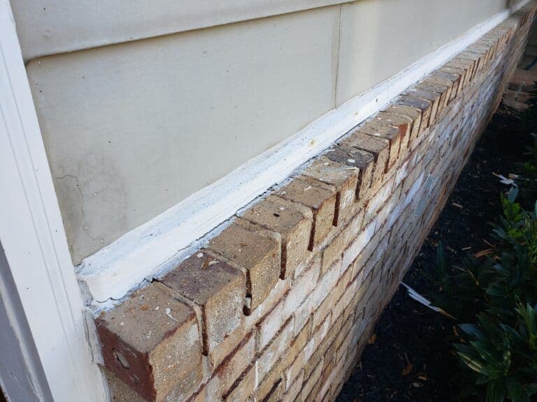 Brick wall façade showing uneven mortar, light weathering, and a thin layer of white paint near the top edge, adjacent to green foliage.