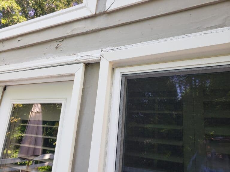 Close-up of a house exterior showing two windows with white frames. The paint on the trim appears chipped and weathered, set against a gray wall.