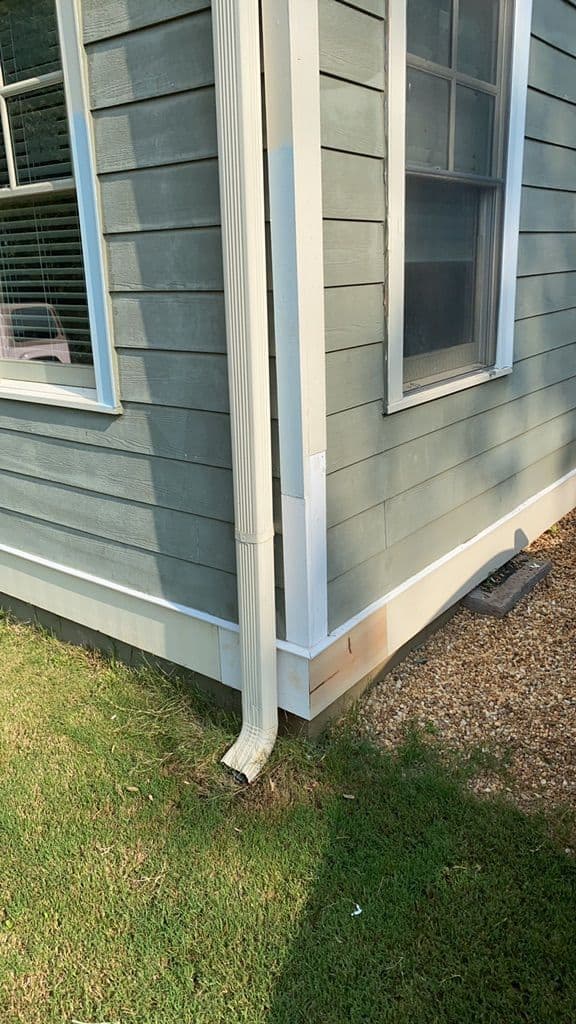 A corner of a light blue house with white trim, featuring a downspout directing water to the grass. Two windows are visible with shadows cast on the lawn.