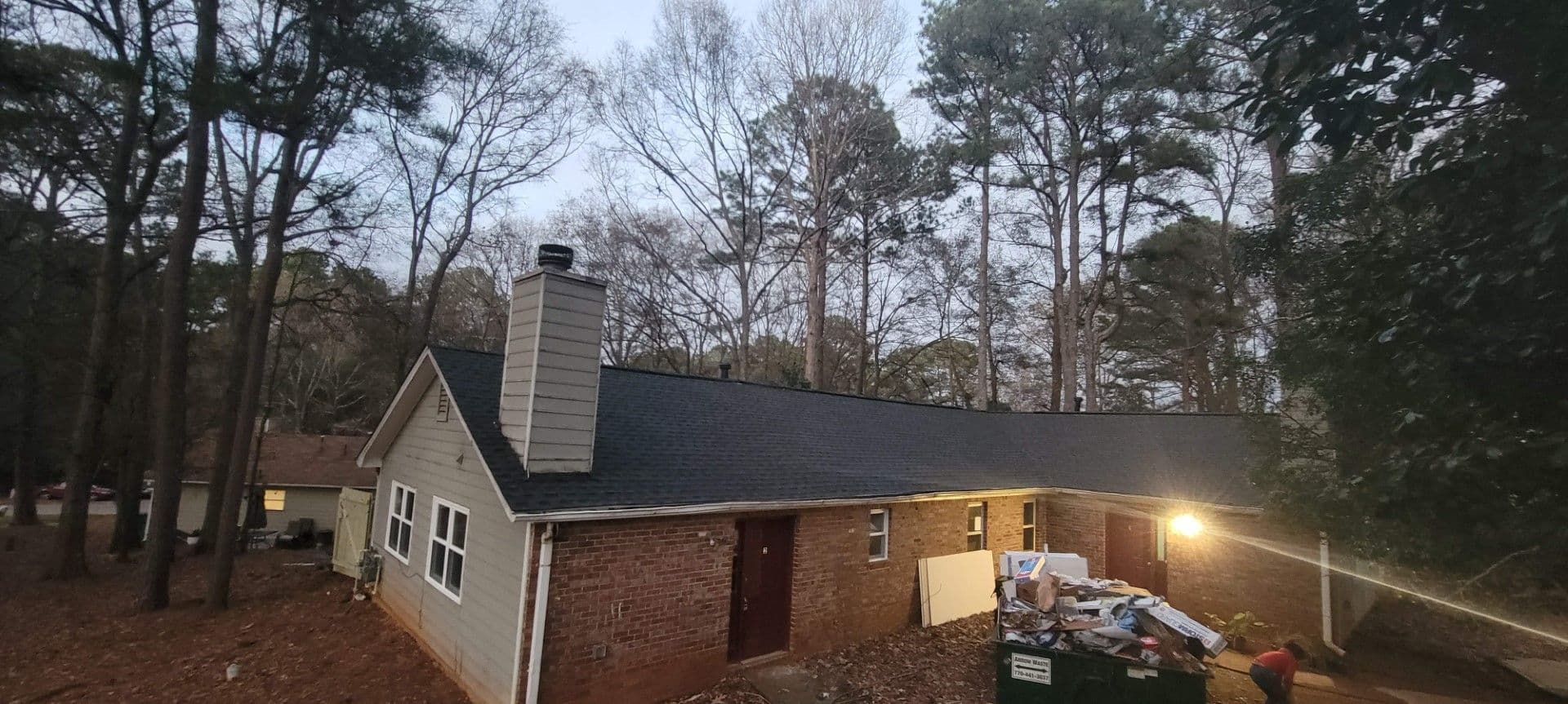 A brick house with a dark roof sits among tall, leafless trees at dusk. A lit porch reveals construction debris and a green dumpster nearby, suggesting renovation.