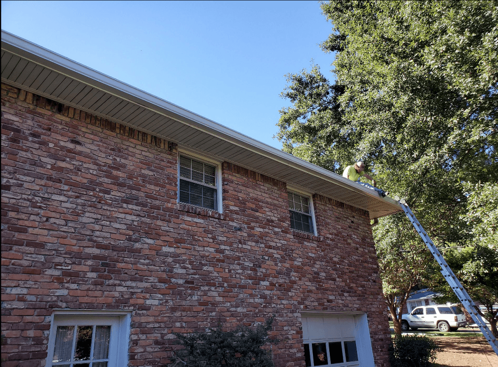 Person in a green shirt stands on a ladder cleaning gutters of a two-story brick house. Sunny day, blue sky, and leafy trees in the background.