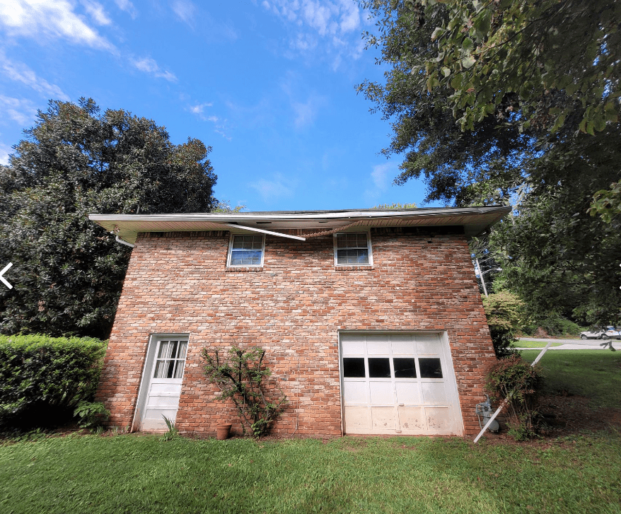 A two-story brick building with a white door and garage, surrounded by lush greenery. Bright blue sky and trees create a serene, natural setting.